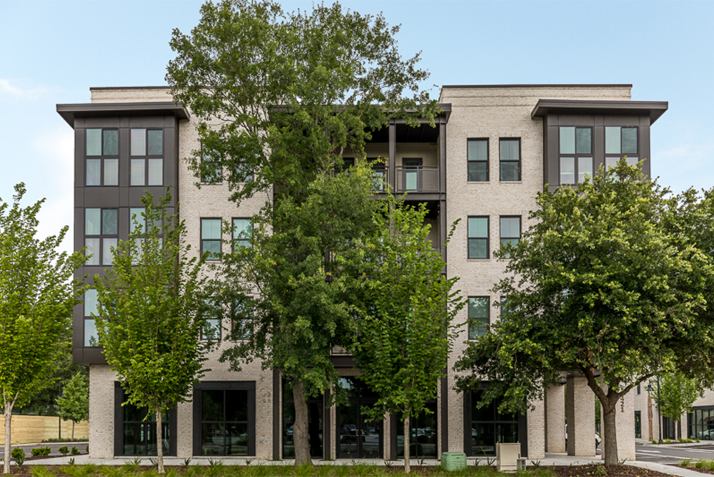 Four-story modern apartment building, crafted by multifamily general contractors, with large windows partially obscured by leafy trees in front, on a clear day.