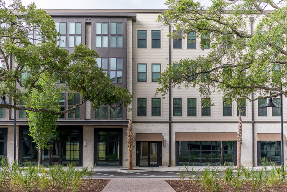 Four-story modern apartment building with large windows and neutral siding, showcasing contemporary multifamily construction, with trees partially obscuring the facade.