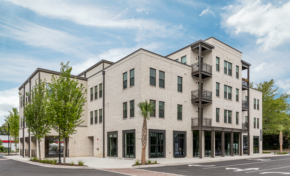 Three-story modern apartment building featuring multifamily construction, large windows, balconies, and ground-floor retail spaces, set on a corner lot with trees and an empty parking lot.
