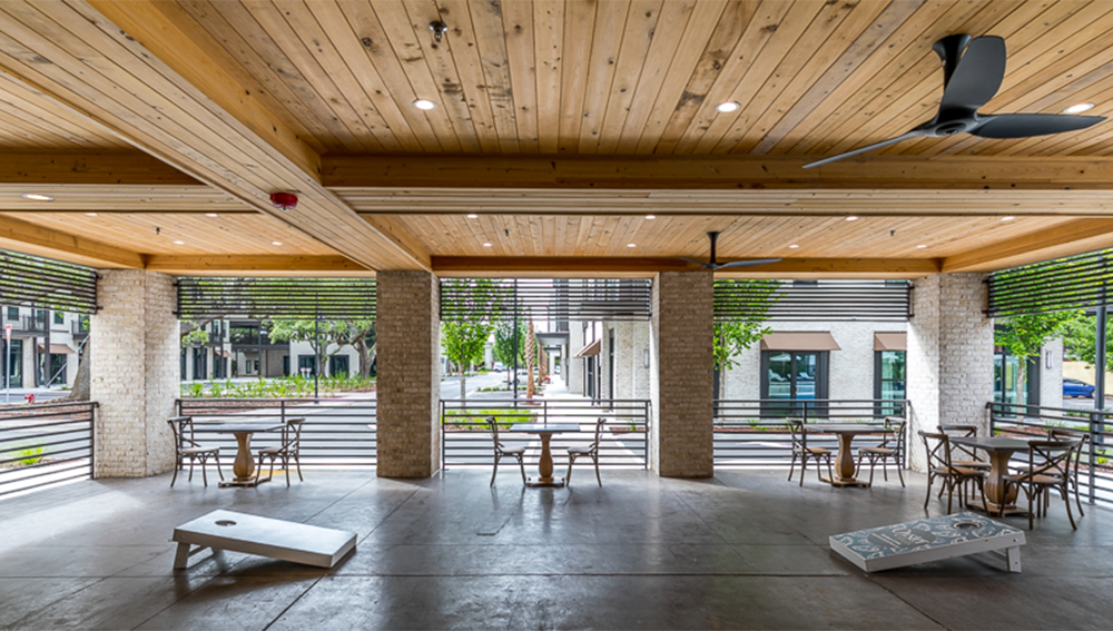 Open-air patio with wooden ceiling, ceiling fan, small tables and chairs, and two cornhole boards; ideal for market rate developments with views of trees and neighboring buildings.
