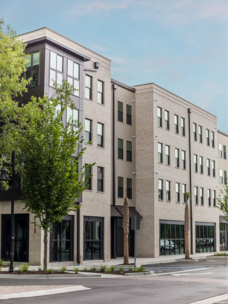 Four-story modern apartment building with large windows, light brick exterior, and street-level retail space, next to a sidewalk with young trees under a clear sky—an ideal example of market rate developments by top multifamily general contractors.
