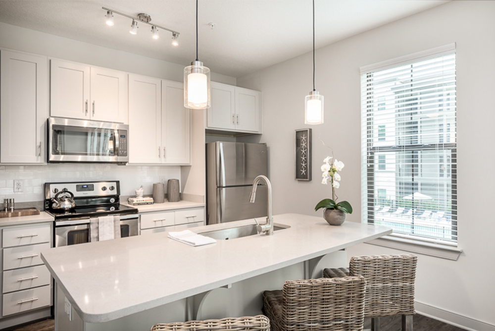 Modern kitchen with white cabinets, stainless steel appliances, island with sink, two woven barstools, pendant lights, and a window with blinds—expertly crafted by leading multifamily general contractors.
