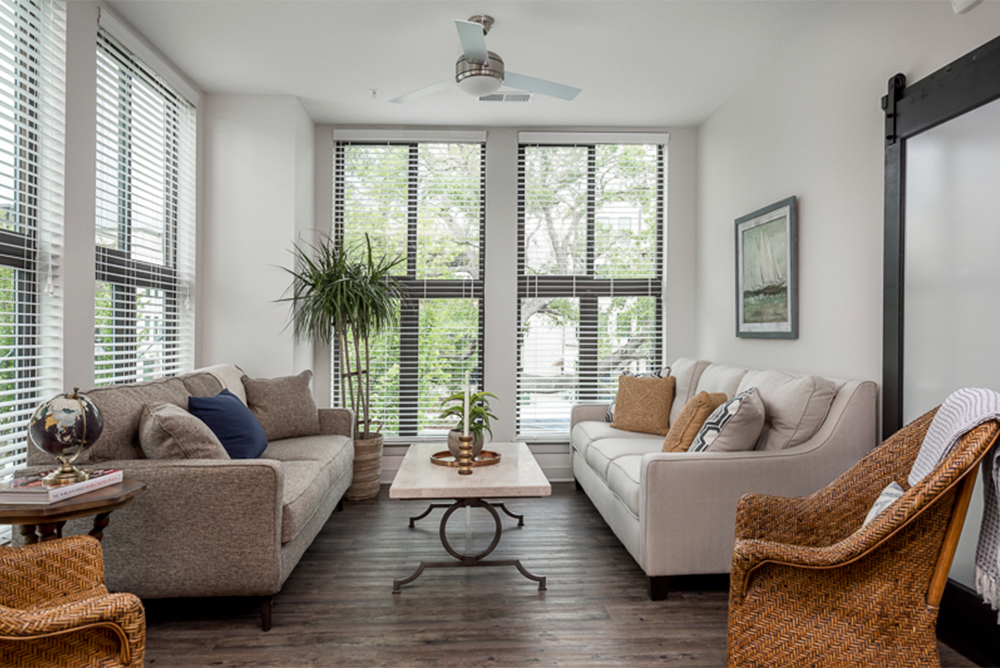 A bright living room with large windows, two sofas, wicker chairs, a coffee table with plants, a potted plant, and a ceiling fan—ideal for market rate developments thoughtfully designed by multifamily general contractors.