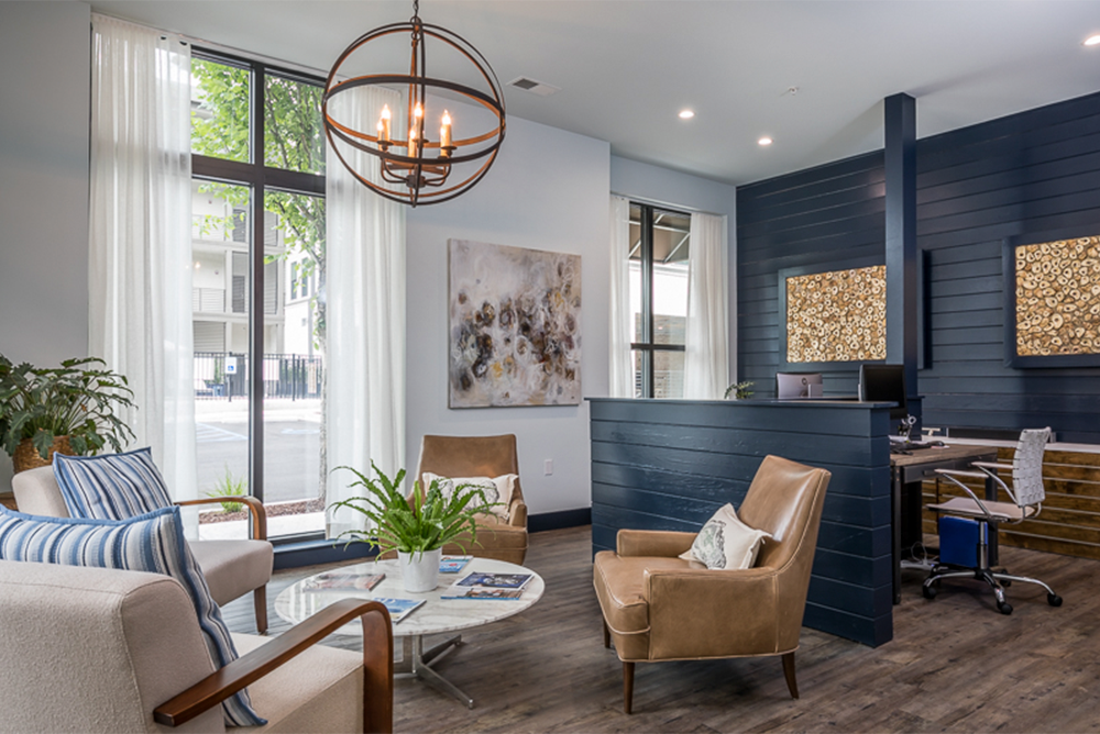 Modern office reception area with blue accent wall, wooden floors, leather and fabric chairs—ideal for multifamily construction or market rate developments—features a round chandelier, large windows, lush plants, and abstract artwork.