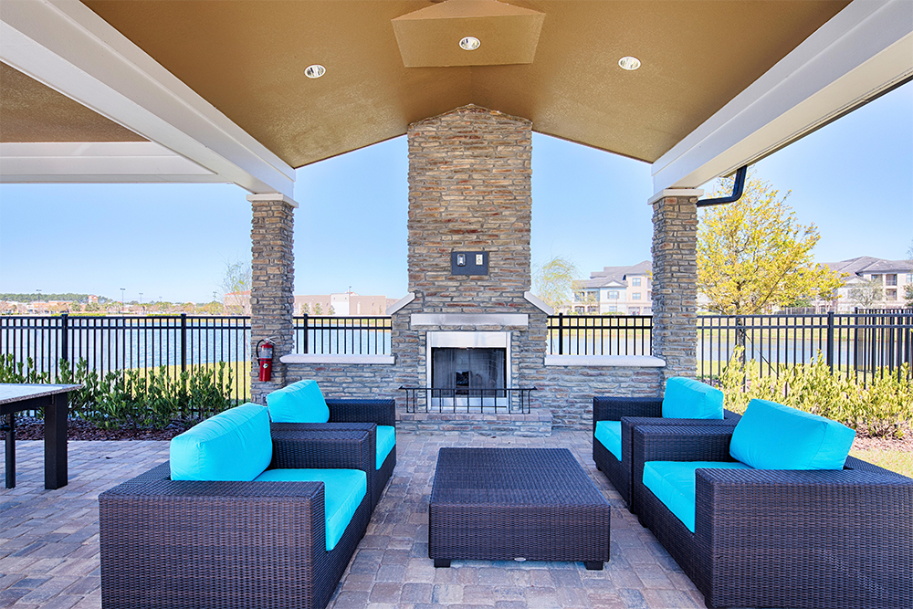 Covered outdoor patio with brown wicker furniture, bright blue cushions, a stone fireplace, and a view of a fenced area with buildings and trees—perfect for modern multifamily construction or land development projects.