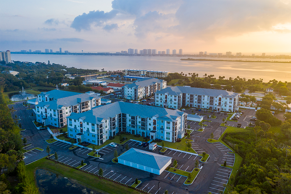 Aerial view of a modern apartment complex showcasing multifamily construction with multiple buildings, parking lots, and landscaped areas near a large body of water at sunrise.