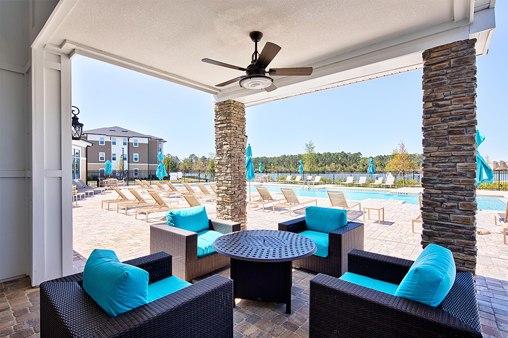 Covered outdoor seating area with wicker chairs and turquoise cushions overlooks a swimming pool—a perfect retreat in this thoughtfully designed space by multifamily general contractors, featuring lounge chairs and umbrellas on a sunny day.