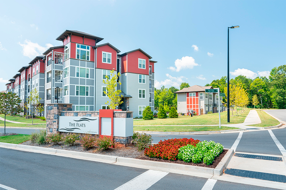 Modern apartment complex called The Flats with red and gray exterior, landscaped grounds, and a street sign visible on a sunny day—an example of market rate developments in multifamily construction.