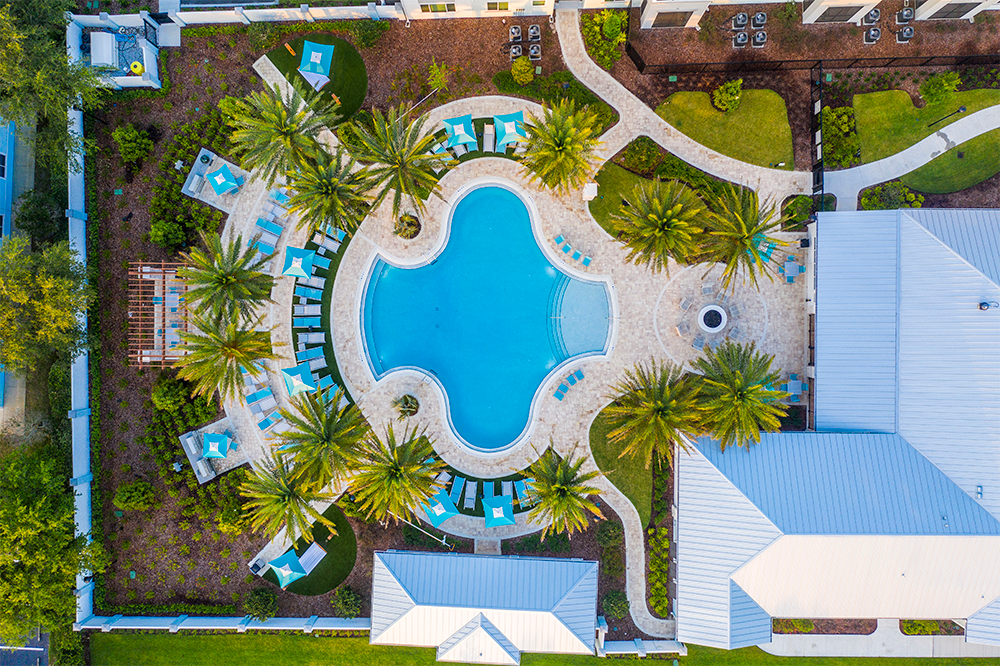 Aerial view of a large, uniquely shaped outdoor swimming pool surrounded by palm trees and lounge chairs, showcasing modern multifamily construction and upscale market rate developments amid nearby buildings.