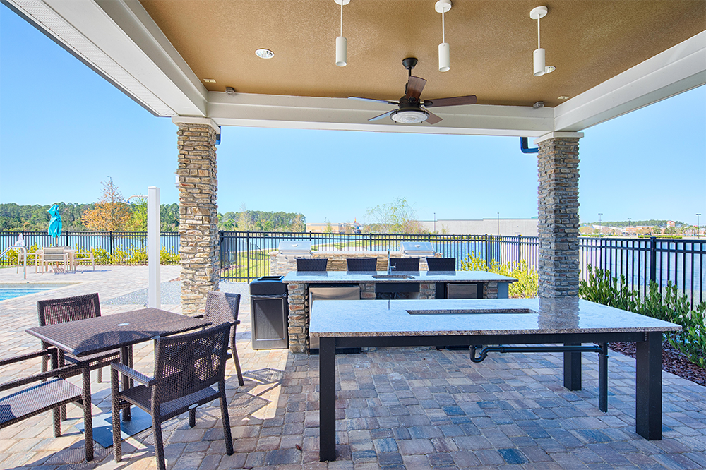 Outdoor covered patio with ceiling fan, stone columns, dining tables, chairs, and a grill area—crafted by multifamily general contractors—overlooking a pool and fenced waterfront view.