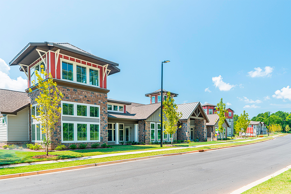 Modern apartment complex with stone and wood facades, large windows, and neatly landscaped grounds—expertly built by multifamily general contractors—along a paved street under a clear blue sky.