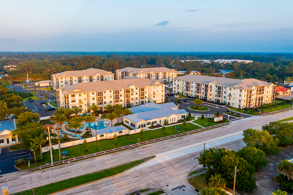 Aerial view of a modern apartment complex, showcasing market rate developments with multiple buildings, a swimming pool, landscaped grounds, and adjacent roads surrounded by trees.
