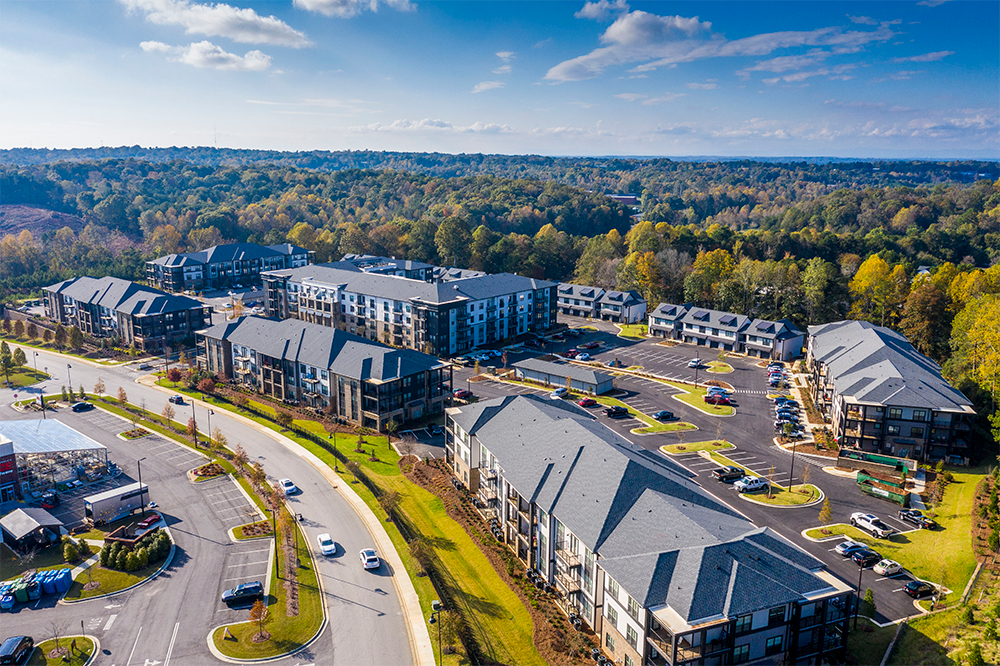 Aerial view of a modern apartment complex, a prime example of multifamily construction, surrounded by trees, parking lots, cars, and landscaped green areas on a sunny day.