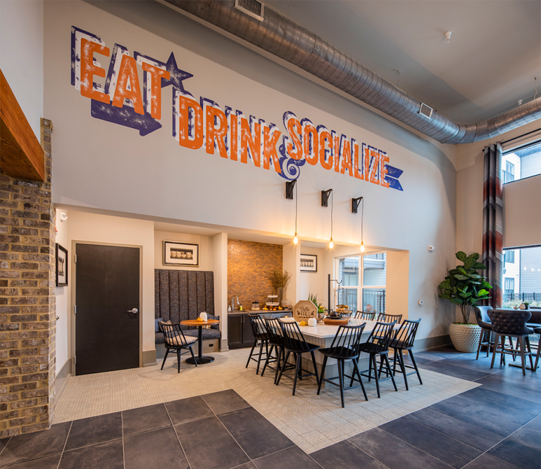 A modern dining area perfect for multifamily construction features a long table, black chairs, decorative plants, and a wall sign reading "EAT DRINK SOCIALIZE" in large, colorful letters.