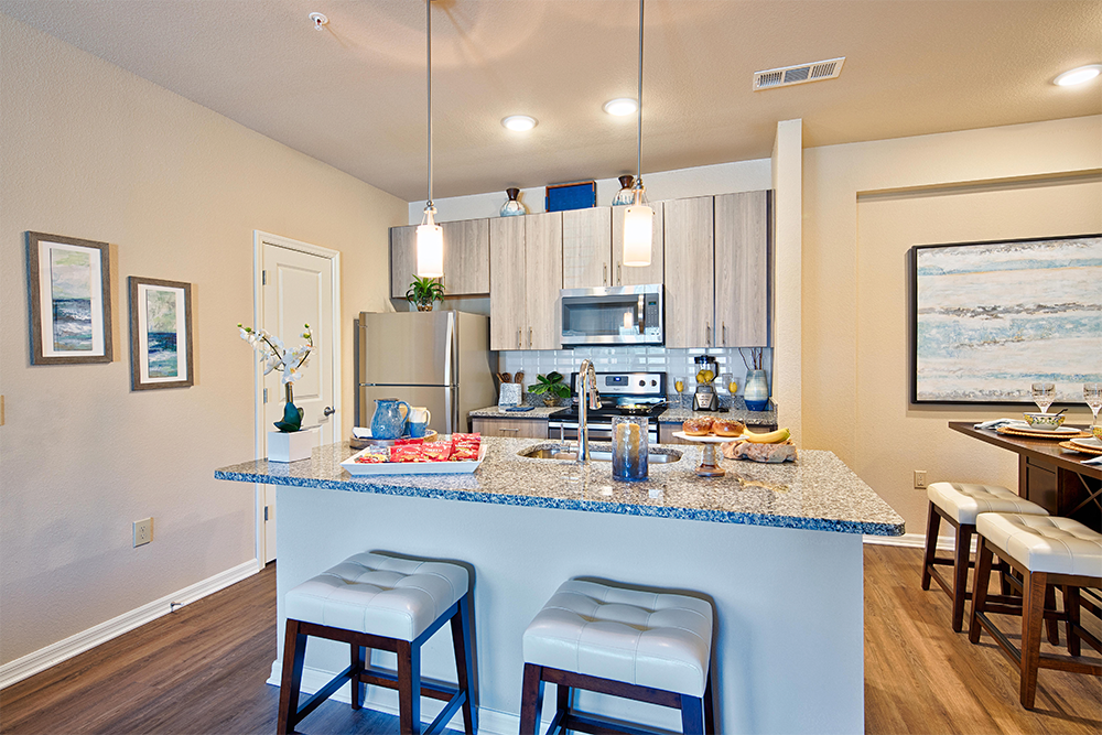 Modern kitchen with granite countertops, stainless steel appliances, and two pendant lights—ideal for market rate developments. Decorated with plants, framed art, and two white stools at the island.