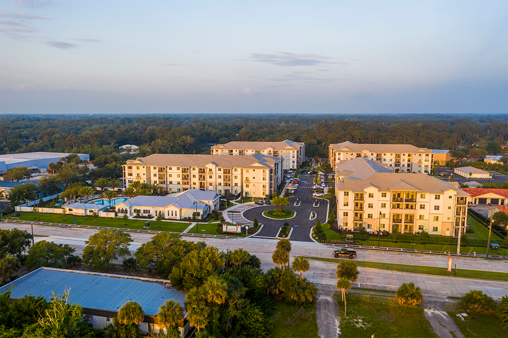 Aerial view of a market rate multifamily construction apartment complex with multiple beige buildings, surrounding greenery, parking areas, and a central roadway.