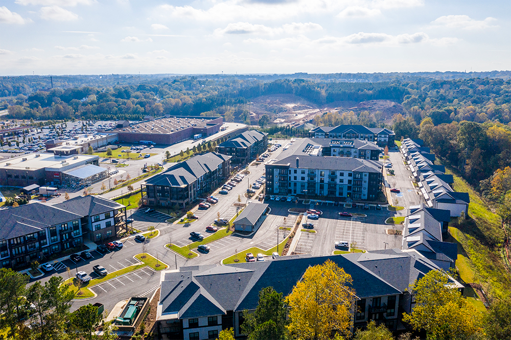 Aerial view of a residential apartment complex, shaped by multifamily construction expertise, surrounded by parking lots, trees, and commercial buildings under a partly cloudy sky.
