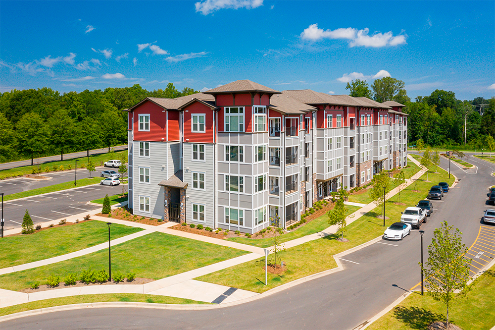 A modern, four-story apartment building with red and gray exterior—an example of market rate developments—surrounded by green lawns, parking lots, and trees under a blue sky.