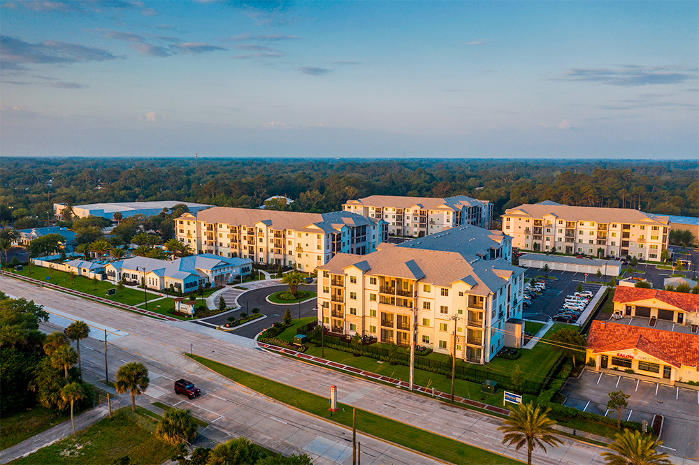 Aerial view of a modern apartment complex built by experienced multifamily general contractors, featuring multiple buildings, parking lots, and landscaped areas beside a wide road, surrounded by trees.