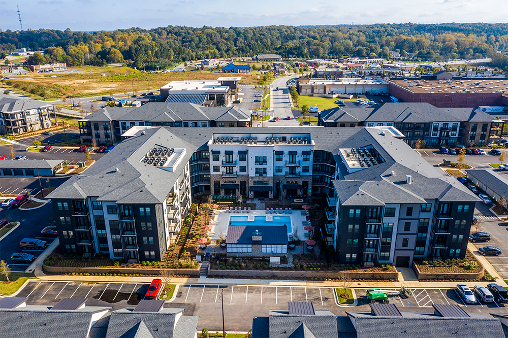 Aerial view of a modern apartment complex showcasing market rate developments with a central swimming pool, surrounded by parking lots, green spaces, and commercial buildings in the background.