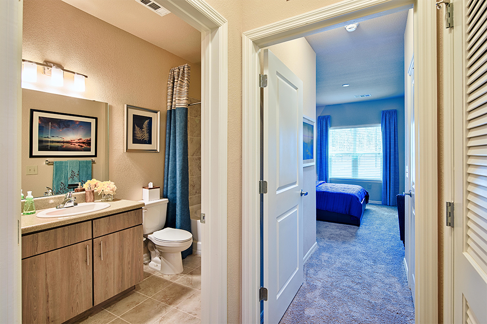 View of a modern bathroom with a vanity, toilet, and wall art, adjacent to a hallway typical of market rate developments, leading to a carpeted bedroom with blue curtains and a neatly made bed.
