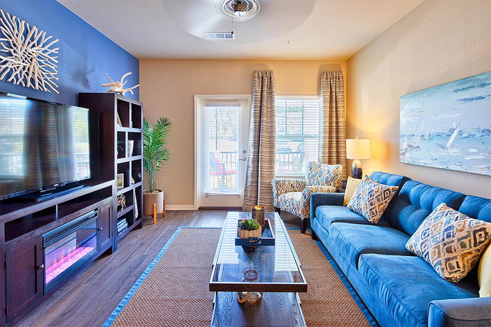Bright living room in market rate developments features a blue accent wall, blue sofa, patterned chairs, glass coffee table, TV stand, large windows with drapes, and modern decor crafted by expert multifamily general contractors.