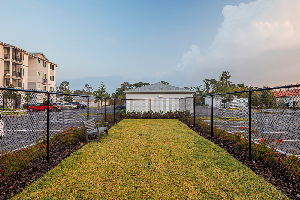 A small, fenced grassy area with a bench, surrounded by parking lots and apartment buildings—a quiet spot amid ongoing multifamily construction and land development, with a white garage visible in the background.