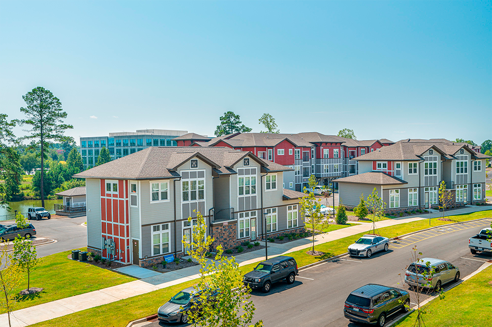 Modern apartment buildings, crafted by multifamily general contractors, line a sunny street with parked cars, green lawns, and trees, while a larger office building stands in the background.