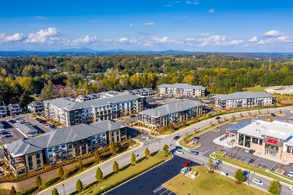 Aerial view of a market rate multifamily construction featuring multiple residential buildings, parking lots, and nearby retail stores, all surrounded by trees and greenery under a blue sky.