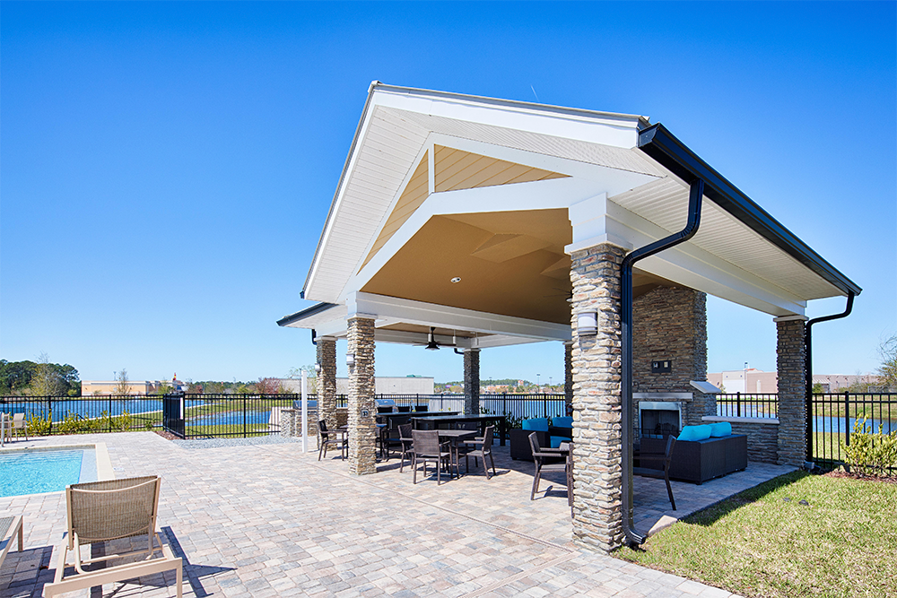 Covered outdoor seating area with stone pillars and a fireplace next to a swimming pool, set on a paved patio under a clear blue sky—ideal for multifamily construction or market rate developments seeking stylish amenities.