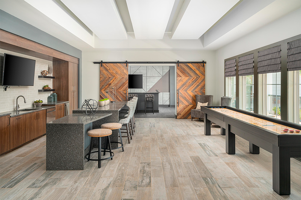 Modern game room with a shuffleboard table, bar seating, wall-mounted TVs, wooden cabinetry, and large windows with blinds—ideal for market rate developments built by expert multifamily general contractors.