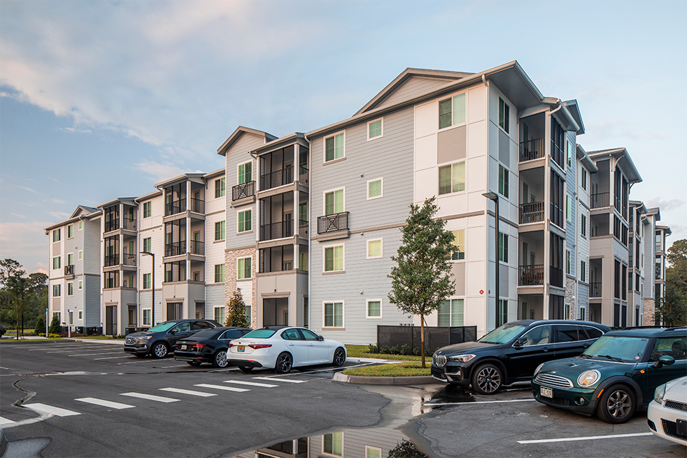 A four-story modern apartment building with balconies, surrounded by parked cars in a lot and some small trees, under a partly cloudy sky—a prime example of market rate developments led by experienced multifamily general contractors.