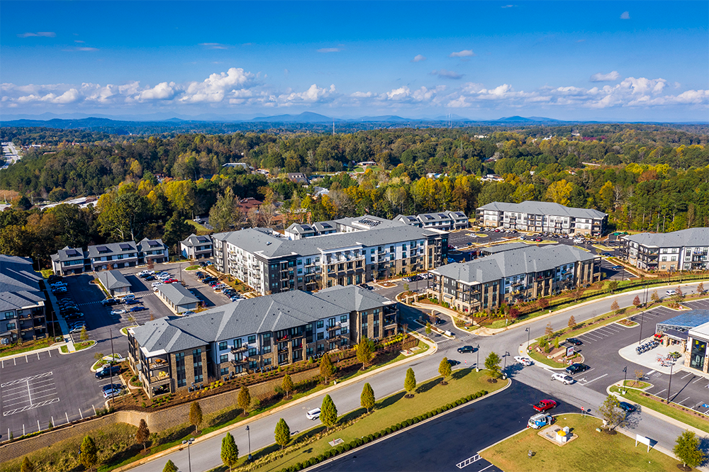 Aerial view of a modern apartment complex, expertly crafted by multifamily general contractors, featuring multiple buildings, parking lots, landscaped areas, and surrounding trees under a blue sky.