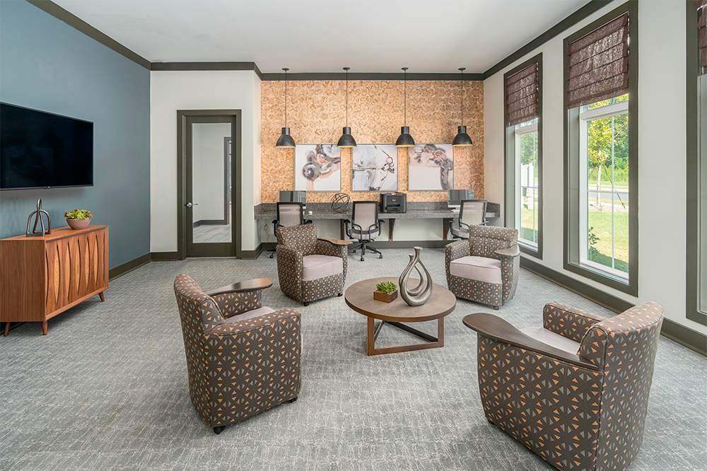 Modern office lounge features four patterned armchairs around a round table, a computer workstation in the back, large windows, and wall art under pendant lights—ideal for multifamily construction or market rate developments.