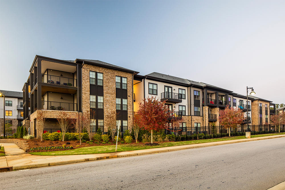Modern three-story apartment building with balconies and brick exterior, showcasing quality multifamily construction, lined with young trees and shrubs along a quiet street.