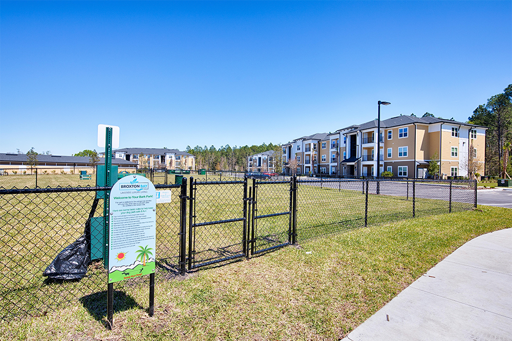 A fenced grassy area with a sign and gate, located near a sidewalk, sits in front of modern apartment buildings—reflecting thoughtful land development and market rate developments—surrounded by trees under a clear blue sky.