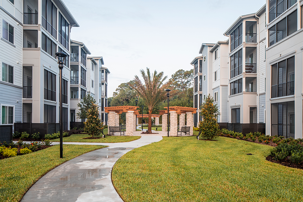 Modern market rate developments feature apartment buildings with balconies surrounding a landscaped courtyard, with a winding sidewalk, green lawn, trees, and a pergola offering outdoor seating.