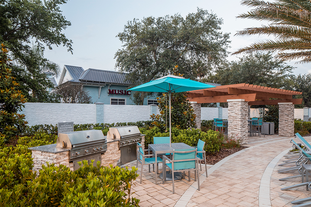 Outdoor patio area with gas grills, dining tables with turquoise chairs, an umbrella, and a wooden pergola surrounded by greenery—perfect for market rate developments seeking inviting amenities.