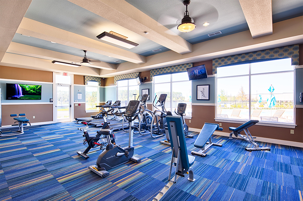 Modern gym in a market rate development, featuring various exercise equipment including stationary bikes, treadmills, and weight benches, with large windows and blue patterned flooring.