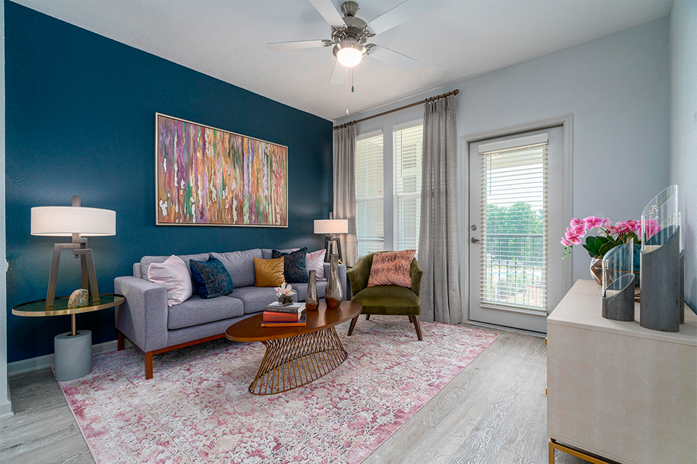 Modern living room with a gray sofa, green chair, wooden coffee table, colorful wall art, pink rug, lamps, and large window with sheer curtains—a stylish space often found in market rate developments or new multifamily construction projects.