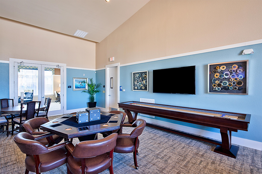A game room designed for market rate developments, featuring a poker table, leather chairs, shuffleboard, wall art, a large TV, and glass doors leading to another room.