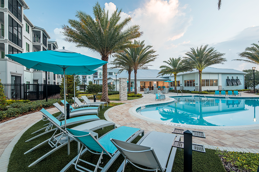 Outdoor swimming pool area with lounge chairs, turquoise umbrellas, palm trees, and modern apartment buildings in the background—a perfect retreat highlighting contemporary multifamily construction.