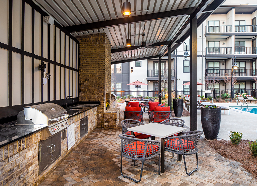 Outdoor patio area with a built-in grill, dining table, chairs, and lounge seating—perfect for market rate developments or multifamily construction—adjacent to a swimming pool and apartment buildings.