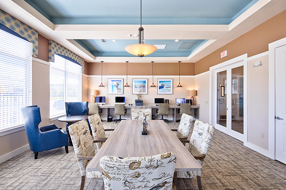 A modern office, ideal for multifamily general contractors or land development teams, features a long wooden table, patterned chairs, blue armchairs, wall art, computers on desks, and large windows letting in natural light.