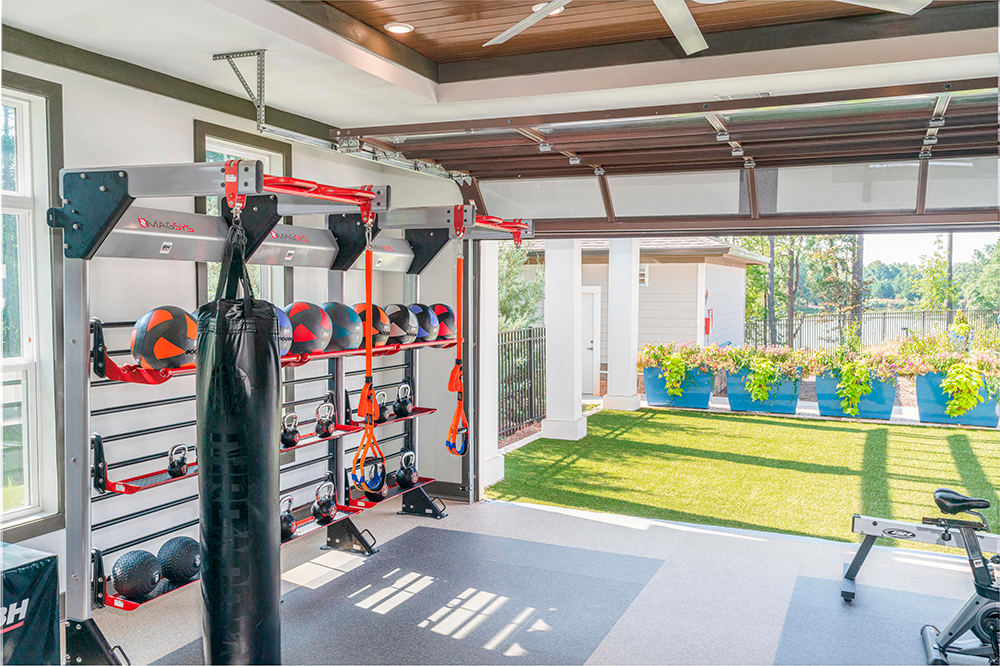 Home gym with exercise equipment, medicine balls, a punching bag, and open garage doors leading to a sunny yard—perfect for residents in market rate developments or those interested in land development and multifamily construction.