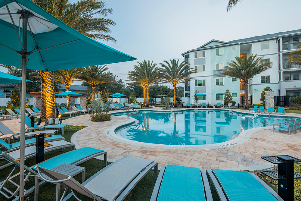 Outdoor swimming pool area with turquoise lounge chairs, umbrellas, palm trees, and a multi-story apartment building in the background at sunset—an inviting amenity found in premium market rate developments.