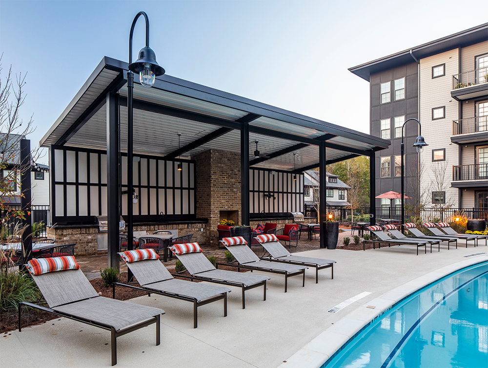 Several poolside lounge chairs with striped pillows face a curved swimming pool. Behind them is a covered seating area, showcasing stylish amenities often found in modern multifamily construction and market rate developments.