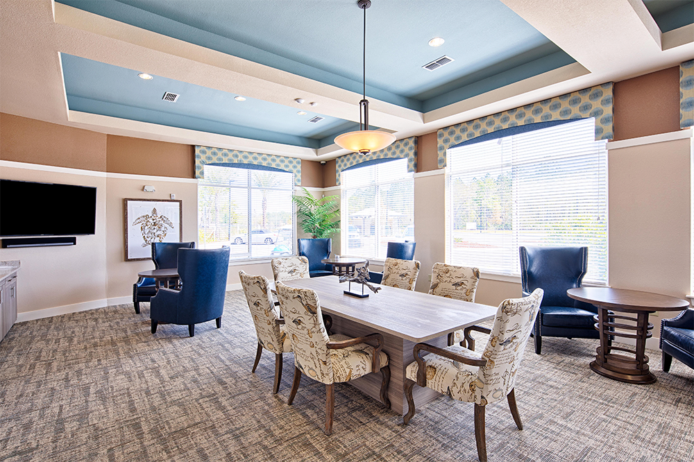 A well-lit meeting room with a rectangular table, eight upholstered chairs, two blue armchairs, large windows, and a flat-screen TV—ideal for land development or multifamily construction project discussions.