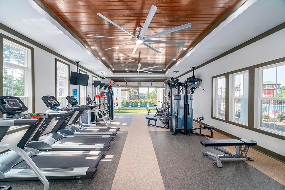 A modern gym in market rate developments, with treadmills, weight machines, benches, and large ceiling fans, featuring windows and views of an outdoor area.