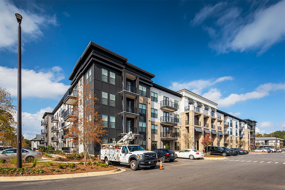 Modern four-story apartment building with balconies, parked cars, and a service truck—an example of quality multifamily construction—stands in the foreground under a partly cloudy sky.